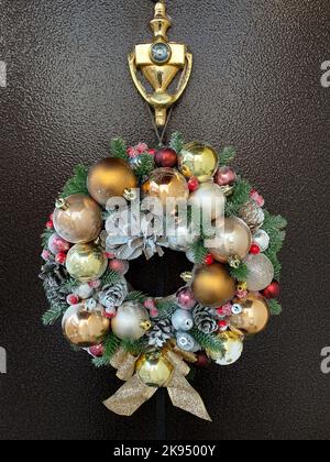 La lussuosa corona decorativa natalizia, fatta a mano con coni di pino e palle dorate, è appesa alla porta anteriore scura della casa. Foto Stock
