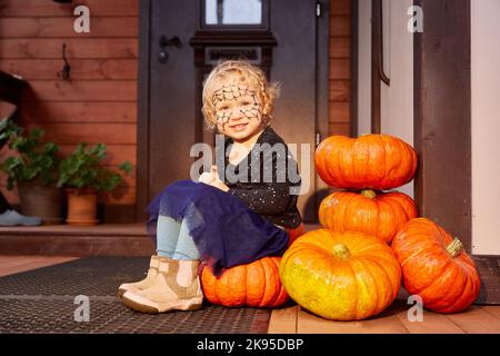 una ragazza sorridente è seduta accanto alle zucche vicino alla porta della casa. arredamento di halloween. Foto Stock