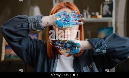 Donna caucasica dai capelli rossi ragazza signora pittore studente di scuola d'arte guardando la macchina fotografica al coperto workshop pittura tenendo mostra Nascondi dietro Foto Stock