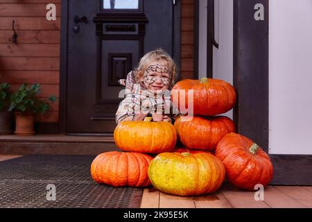 una ragazza sorridente è seduta accanto alle zucche vicino alla porta della casa. arredamento di halloween. Foto Stock