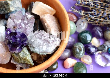 Pietre semi-preziose in una ciotola di legno con bouquet di lavanda asciutto. Cristalli di druso di ametisti e ametisti, quarzo rosa, agato, apatite, aventurina, turq Foto Stock