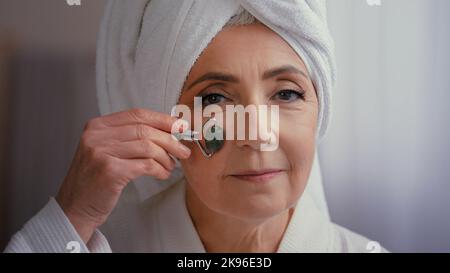 60s anziana di mezza età donna caucasica matura con asciugamano sulla testa in bagno massaggio mattutino viso con rullo di pietra anziana bella donna cura della pelle Foto Stock