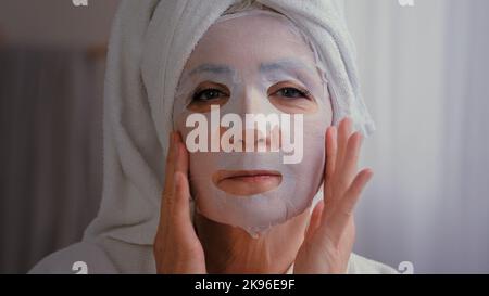 Ritratto in bagno vecchia donna causiana anziana maturo wrap tovagliolo sulla testa nonna donna applicare skincare maschera in tessuto sulla faccia femminile trattamento viso Foto Stock