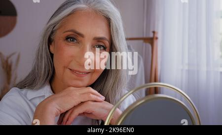Senior grigio-capelli più anziano 60s donna 50s signora granny con rugoso bel viso di invecchiamento con perfetta pelle liscia guardando specchio riflessione toccando Foto Stock