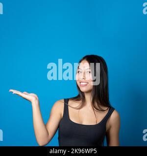 Giovane donna alla moda multirazziale con grande sorriso tenendo e presentando spazio copia sulla sua palma isolato su sfondo blu Foto Stock