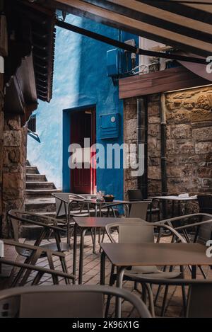 Un colpo verticale di vicolo di bar in villaggio con facciata blu, porta rossa, sedie di plastica grigia e tavoli sotto tende Foto Stock