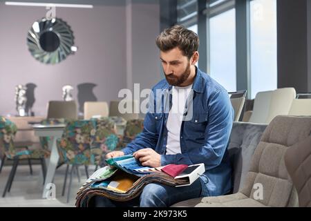 Uomo bearded scegliendo il colore e la texture del tessuto della tappezzeria da vari campioni nel deposito della mobilia. Vista laterale del cliente maschio che tocca i tessuti, facendo scelta, mentre si siede sul divano. Concetto di design. Foto Stock