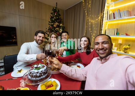 Videochiamata per Natale, vari amici che parlano su uno smartphone e scattano una foto congiunta selfie seduti a casa al tavolo delle feste vicino all'albero di Natale, vacanze di Capodanno. Foto Stock