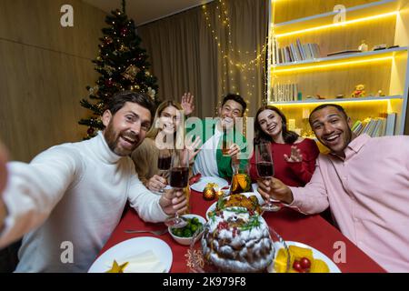 Videochiamata per Natale, vari amici che parlano su uno smartphone e scattano una foto congiunta selfie seduti a casa al tavolo delle feste vicino all'albero di Natale, vacanze di Capodanno. Foto Stock