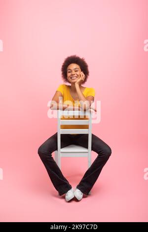 Sorridente giovane donna gara mista con capelli ricci casual si siede sulla sedia, isolato su sfondo rosa Foto Stock