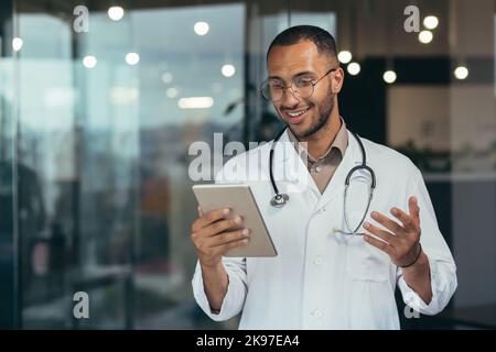 Un giovane bel medico ispanico maschile sta avendo una consultazione online con un paziente. In piedi in ospedale con uno stetoscopio, tenendo in mano una tavoletta, spiegando con le mani. Foto Stock