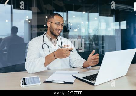 Ricevimento online presso l'ospedale. Giovane e bello medico ispanico che parla con un paziente durante una videochiamata da un portatile. Si siede in ufficio alla scrivania, parla con la fotocamera, spiega e fa le sue mani con le onde Foto Stock