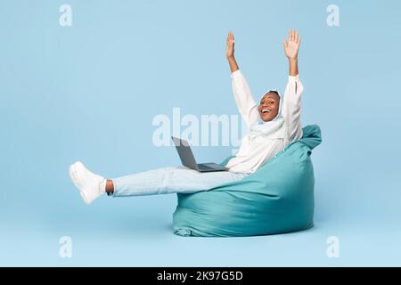 Donna musulmana afro-americana overjoyed seduta in sedia di beanbag con il laptop e alzando le mani in su, ha ottenuto le notizie grandi Foto Stock