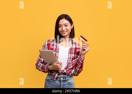 Concetto di shopping online. Donna cinese felice che tiene il tablet digitale e mostra la carta di credito, ordinando gli acquisti Foto Stock