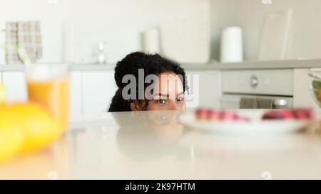 Giovane donna nera che sceglie tra pasto sano e non sano, sbirciando fuori dal tavolo e guardando la ciambella dolce Foto Stock