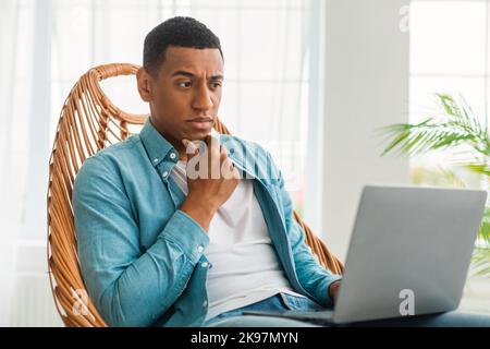 Un giovane uomo nero molto bello e pensivo in un look casual al computer portatile, si siede su una sedia negli interni luminosi della stanza Foto Stock