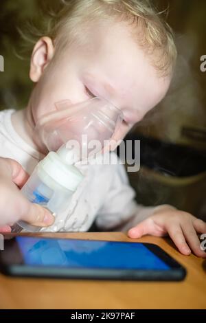 Piccolo bambino carino in maschera di un inalatore. Procedura di inalazione a casa. Bambino che prende la terapia respiratoria con il nebulizzatore. Guardare i cartoni animati sul Foto Stock