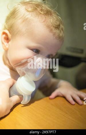 Piccolo bambino carino in maschera di un inalatore. Procedura di inalazione a casa. Bambino che prende la terapia respiratoria con il nebulizzatore. Guardare i cartoni animati sul Foto Stock