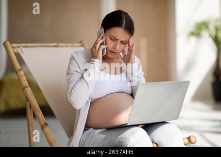Donna incinta stressata che usa il laptop e che parla sul cellulare a casa Foto Stock