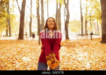 Ritratto orizzontale di donna sorridente in intimo maglione a maglia rosso che getta su bouquet di foglie gialle su fondo foresta dorata. Giovane donna felice Foto Stock