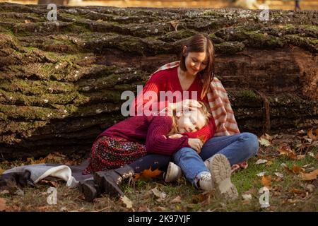 Ritratto della bambina anziana della scuola sdraiata sulle gambe della madre amorevole in abbracci, fondo di tronco d'albero abbattuto. Donna felice pat sulla testa figlia seduta Foto Stock