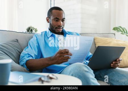 Concetto di documentazione. Ritratto dell'uomo nero che lavora a casa, leggendo carta o documento finanziario, seduto sul divano Foto Stock
