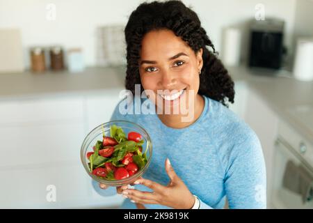 Donna afro-americana felice in abbigliamento sportivo mangiare insalata sana dopo l'allenamento domestico, seduta in cucina e sorridente Foto Stock