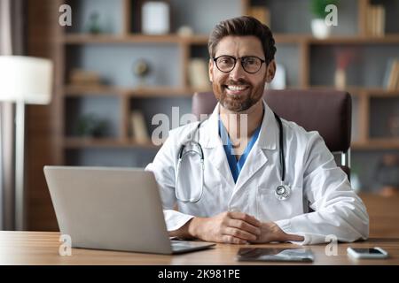 Bel medico maschile in uniforme seduto alla scrivania con computer portatile sul posto di lavoro Foto Stock