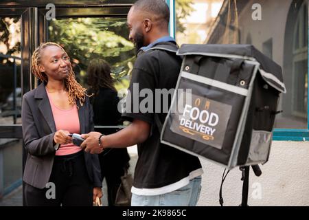 Pagamento senza contatto del terminale POS per la consegna degli ordini di cibo, donna che paga il pasto da asporto con carta di credito. Corriere Africano-americano che consegna il pranzo del ristorante al dipendente dell'ufficio Foto Stock