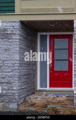 Una parete esterna in mattoni grigi di stile moderno di una casa con un'entrata ad una luminosa porta rossa. Ci sono tre piccole finestre nella porta. Foto Stock