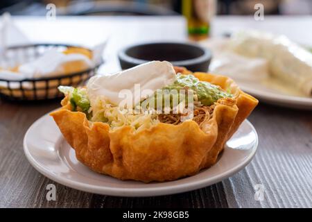 Una croccante ciotola messicana di taco fatta con una tortilla rotonda cotta ripiena di fagioli, mais, manzo, panna acida, guacamole, avocado, lime, cipolle, pomodori. Foto Stock