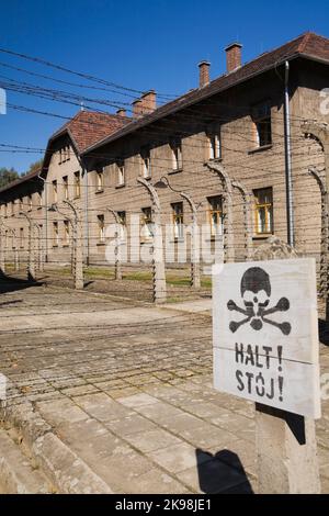 Stop segnale di avvertimento di fronte a una recinzione elettrificata di filo metallico e edificio all'interno dell'ex campo di concentramento nazista di Auschwitz i, Polonia. Foto Stock