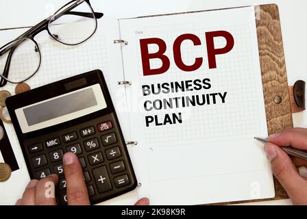 Su uno sfondo grigio chiaro si trovano una penna, un notebook verde e un biglietto bianco con il testo BCP Business Continuity Plan. Concetto aziendale. Foto Stock