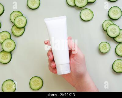 Tubo bianco senza marchio per spremere la crema in mano dell'uomo. Bottiglia per marcare a caldo e fette di cetriolo fresco su sfondo verde chiaro. Vista dall'alto, mockup, template. Foto Stock