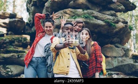 Felice uomo afro-americano turista sta prendendo selfie con gli amici nella foresta vicino a enormi rocce utilizzando smartphone, la gente sta facendo volti divertenti e mostrando gesti di mano fredda. Foto Stock