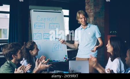 Il team leader maschile sta facendo un rapporto sulla lavagna parlando e indicando la tabella, i membri del team stanno ascoltando e poi battendo le mani. Business, educazione aziendale e concetto di persone. Foto Stock
