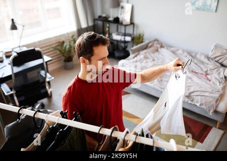 Giovane bell'uomo che sceglie gli abiti da indossare al mattino mentre si trova in piedi nella sua stanza Foto Stock