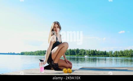 Bella, atletica giovane donna bionda, allenatore, istruttore, stretching, facendo diversi esercizi. Lago, fiume, cielo blu e foresta sullo sfondo, estate giorno di sole. Foto di alta qualità Foto Stock