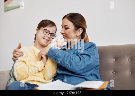 Ritratto di ragazza felice adolescente con sindrome di Down cocdling con la madre mentre si siede sul divano a casa Foto Stock