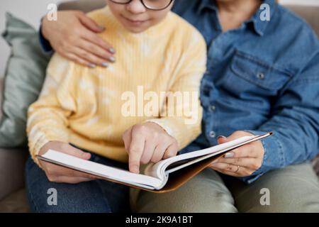 Primo piano di una giovane ragazza con sindrome di Down lettura libro con amorevole madre a casa Foto Stock