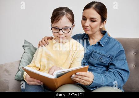 Ritratto di ragazza teen con sindrome di Down lettura libro con amorevole madre a casa Foto Stock