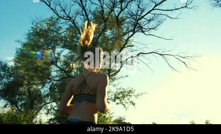 Bella, alta, atletica bionda giovane, ragazza mette le cuffie e inizia a correre, in cima e shorts fa una corsa, lungo il sentiero prato, vicino alla foresta, al tramonto, ai raggi del sole. Foto di alta qualità Foto Stock