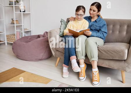 Ritratto completo di ragazza adolescente con sindrome di Down lettura libro con la madre amorevole a casa Foto Stock