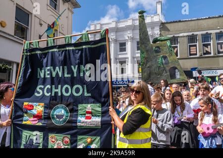 Gli adulti e i bambini della scuola di Newlyn suonano gli strumenti che sfilano con la loro enorme scultura di una miniera di stagno Cornish attraverso Penazance sul co Foto Stock