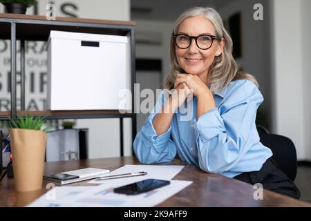 Con successo la testa della donna anziana di mezza età si siede a un tavolo e guarda la telecamera con le mani afferrate in un lucchetto, facendo funzionare un successo Foto Stock