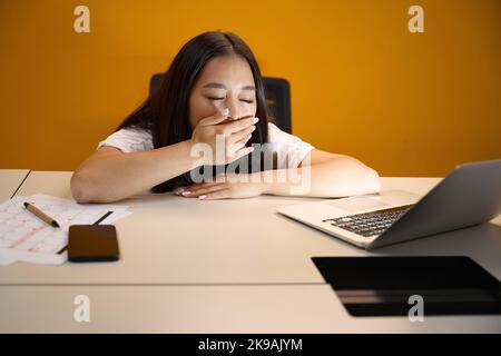 Donna asiatica addormentata sta coprendo la bocca con la mano sul posto di lavoro Foto Stock