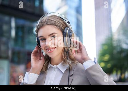 Donna felice ascolta la musica in cuffia mentre cammina nel centro città, sorridendo Foto Stock