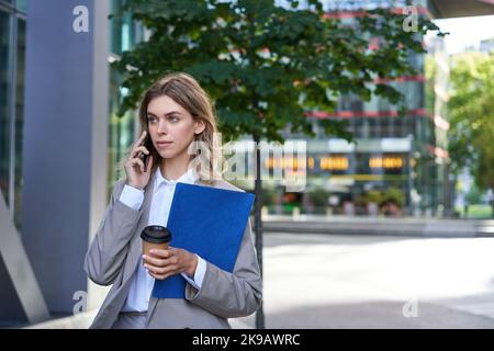 Occupato donna d'affari beve caffè, parla sul telefono cellulare, tiene documenti per il lavoro, passeggiate nel centro della città vicino ufficio Foto Stock
