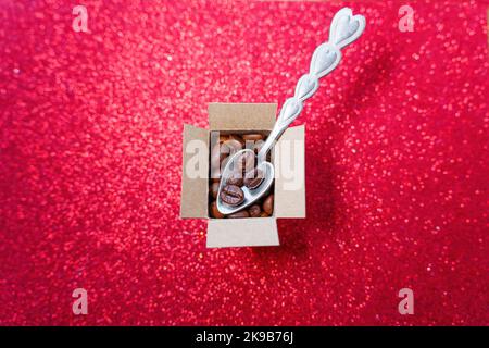 Cucchiaio a forma di cuore collocato in una piccola scatola di cartone piena di chicchi di caffè tostati e collocato su uno sfondo rosso brillante. Foto Stock