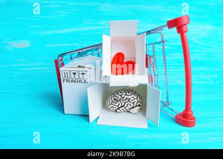Carrello rovesciato con piccole scatole di spedizione con cervello umano e simbolo del cuore isolato su blu. Concetto creativo di shopping terapia. Foto Stock